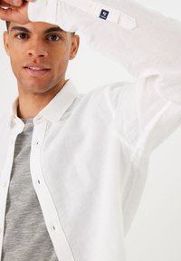 Camisa blanca de lino abotonada con una textura sutil, superpuesta sobre una camiseta gris a rayas. Incluye una etiqueta de marca en la manga.