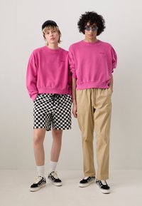 Dos personas llevan sudaderas rosas a juego. Una lleva pantalones cortos de cuadros en blanco y negro; la otra lleva pantalones caqui. Ambas calzan zapatillas deportivas informales.