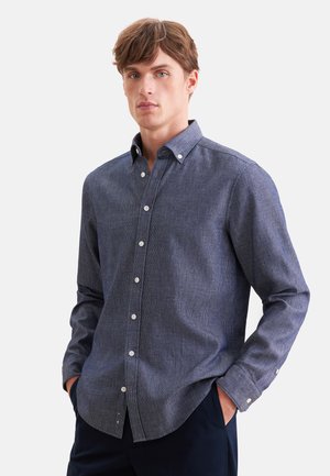 Jeune homme portant une chemise bleue à manches longues boutonée, les mains dans les poches, debout devant un fond blanc uni.