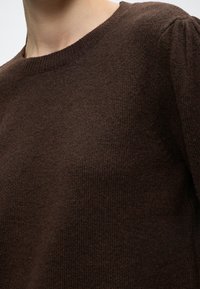 Gros plan d'une personne portant un pull en tricot marron foncé avec un col rond et de subtiles fronces aux épaules.