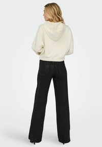 Krémová mikina s kapucí na šňůrku a elastickým lemem, spolu s černými džíny s širokými nohavicemi a vysokým pasem. Hladká textura, volný design.