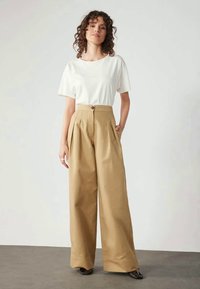 Calças largas bege com pregas e fecho de botão, combinadas com uma t-shirt justa de manga curta branca. Tecido suave, design minimalista.