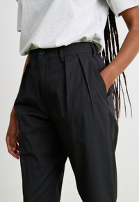 Pantalons noir taille haute avec plis, en tissu lisse. Comprend des poches latérales et une fermeture à bouton à la taille.