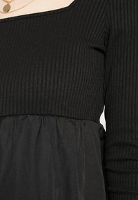 Vestido preto com um corpete canelado e decote quadrado, transicionando para uma saia transparente e drapeada. Tecido texturizado com design em camadas.