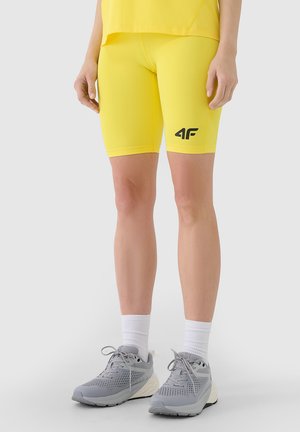 Osoba nosząca jasne żółte spodenki rowerowe z czarnym logo "4F", białe skarpetki i szare buty sportowe, stojąca na jednolitym tle.
