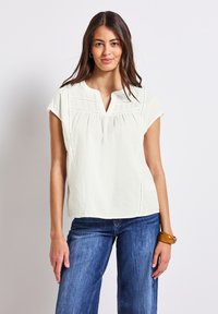 Blusa blanca de manga corta con escote en V, detalles fruncidos y sutiles acentos de costura. Fabricada en una tela suave y ligera.