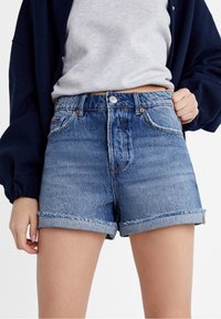 Modelo vistiendo unos pantalones cortos de denim azul con dobladillo enrollado, una camiseta gris metida por dentro y una chaqueta navy oscura, todo ello sobre un fondo blanco.