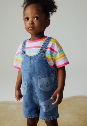Ungt barn med krøllet hår iført farverig stribet skjorte og denim-dragter dekoreret med katte ansigter, stående og kigger let opad.