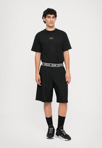 Μαύρη μπλούζα με μικρό λογότυπο, συνδυασμένη με μαύρες βερμούδες που έχουν λευκή ζώνη με λογότυπο. Το μοντέλο φοράει μαύρες κάλτσες και αθλητικά παπούτσια.