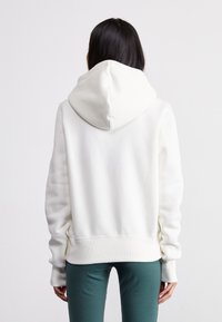 Hoodie branco com capuz espaçoso, punhos e bainha canelados. Textura suave, design casual e ajuste descontraído. Usado com leggings justas em tom de teal.