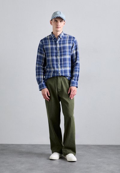 Μπλε καρό πουκάμισο με κουμπιά, ελαιόχρωμο παντελόνι και λευκά αθλητικά παπούτσια. Φοριέται ένα ανοιχτό μπλε καπέλο. Παρουσιάζει απλά σχέδια και χαλαρή εφαρμογή.