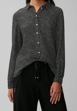 Vrouw draagt een zwart overhemd met lange mouwen en witte polkadots, gecombineerd met zwarte broek met aantrekkoorden en ritszakken.