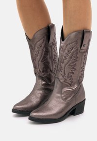Botas de cowboy de bronce metálico con puntas puntiagudas, costuras decorativas y tacones ligeramente elevados. Fabricadas en cuero texturizado con un acabado suave.
