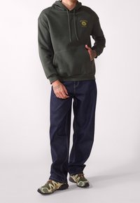 Hombre con sudadera con capucha verde oscuro con texto "Exclusive Local Flavor", jeans azul oscuro y zapatos deportivos verdes y beige, de pie contra un fondo blanco.