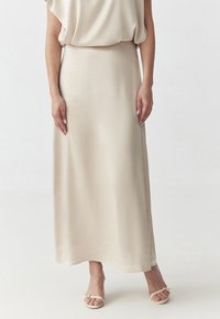 Jupe maxi satin beige avec une texture lisse, un design fluide et une taille ajustée. Associée à des sandales simples de couleur claire.