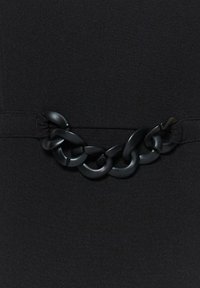 Détail de maillon de chaîne en plastique noir mat sur fond de tissu noir, attaché à de larges bandes élastiques de chaque côté.