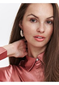 Różowa jedwabna koszula z wywijanym kołnierzykiem i ozdobnym guzikiem, zestawiona z perłowymi kolczykami w kształcie obrączek. Modelka ma długie włosy i neutralny kolor ust.