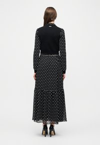 Γυναίκα με μακριά μαλλιά που στέκεται, ντυμένη με μακρυμάνικο φόρεμα που έχει μαύρα και λευκά πουά, μαύρο γιλέκο χωρίς μανίκια και μαύρα τακούνια.