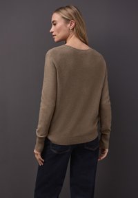 Pull kaki avec une finition texturée, poignets et ourlet côtelés. Porté avec un jean taille haute bleu foncé. Vue de dos sur un fond gris.