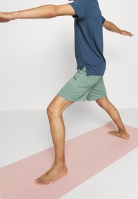 Sportovní oblečení s modrým tričkem a zelenými pruhovanými šortkami, stojící na růžové jógové podložce, přičemž prezentuje lehký design a prodyšnou tkaninu.