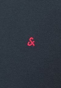 Tmavě modrý textil s texturovaným tkaním, s malým červeným vyšitým logem v pravém dolním rohu.