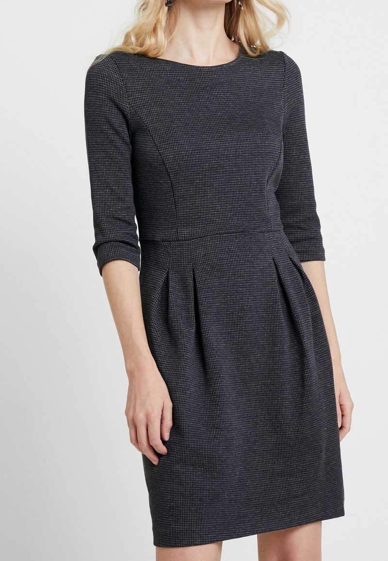 Femme portant une robe ajustée gris foncé avec des manches trois-quarts et des plis sous la taille.