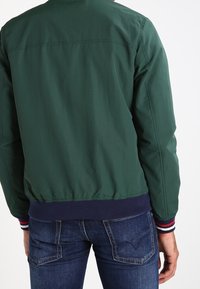 Muž v tmavě zelené bomber bundě s tmavě modrými žebrovanými manžetami a lemem, doplněný modrými džínami, zobrazený zezadu.