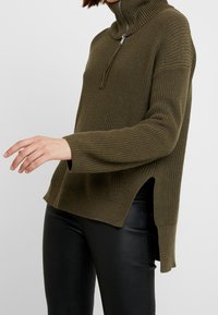 Maglione a collo alto lavorato a maglia verde oliva con zip frontale, spacco laterale e polsini a coste. Tessuto testurizzato e vestibilità ampia.