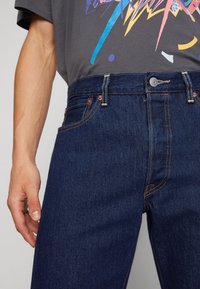 Dunkelblaue Denim-Jeans mit Kontrastnähten, Fünf-Taschen-Design und Knopfverschluss. Kombiniert mit einem grauen Graphic-T-Shirt darüber.