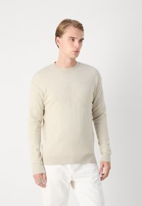 Maglione beige chiaro con un collo rotondo a coste e maniche lunghe. Presenta un logo 'CK' in rilievo sul davanti. Texture a maglia liscia.