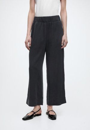 Ženska, oblečena v široke črne hlače z elastičnim pasom in črne balerinke s konicami in zanko za peto, stoji pred enobarvnim ozadjem.