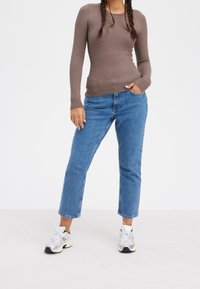 Top lungo a maniche ribatte di colore marrone abbinato a jeans dritti blu chiaro e sneakers grigie. L'outfit è casual, con una finitura tessurizzata sulla parte superiore.