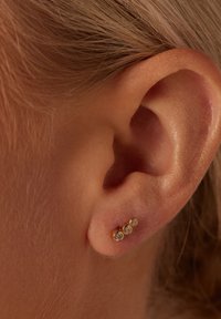 Goldfarbene Ohrringe mit drei klaren, runden Edelsteinen, die in einer Reihe angeordnet sind und am unteren Teil eines Ohres befestigt sind. Glatte Metalloberfläche, dezenter Glanz.