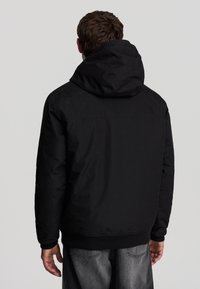 Giacca nera con cappuccio, dal tessuto morbido e liscio, con polsini e orlo a costine. Presenta un design minimalista senza loghi o decorazioni visibili.