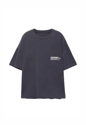 T-shirt in cotone oversized di colore scuro con maniche corte. La parte anteriore presenta testo bianco nella zona del petto a sinistra. Design semplice e privo di fantasie.
