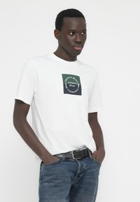 Λευκό βαμβακερό T-shirt με τετράγωνο γραφικό που περιλαμβάνει σκοτεινές πράσινες και μπλε ενότητες. Το κείμενο περιλαμβάνει τη φράση «JACK & JONES» σε κυκλικό σχέδιο.