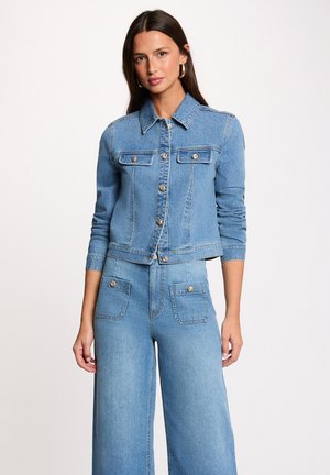 Donna che indossa una giacca di jeans azzurro chiaro con bottoni dorati e jeans a gamba larga a vita alta abbinati, in piedi davanti a uno sfondo semplice.