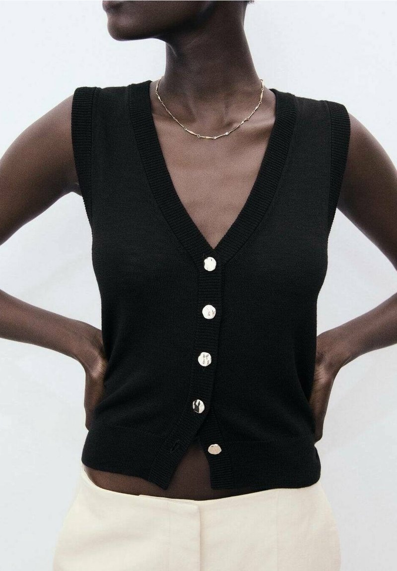 Personne à la peau foncée portant un gilet noir sans manches boutonné et un fin collier chaîne en or, mains posées sur les hanches, pantalon beige visible.