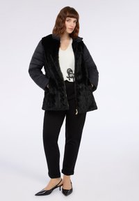 Vrouw in een zwarte gewatteerde jas met nepbont, witte blouse met zwart ontwerp, zwarte broek en zwarte spitse slingback hielen, staand met handen in haar zakken.