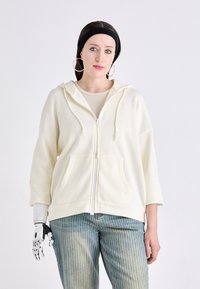 Lys kremfarget hettegenser med glidelås, frontlomme, snor og løs passform, kombinert med blåstripete jeans. Inkluderer en proteseshånd.