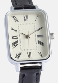 Reloj rectangular en tono plateado con esfera color crema, números romanos, correa de piel negra y una corona plateada en el lateral. Manecillas de horas y minutos sencillas.