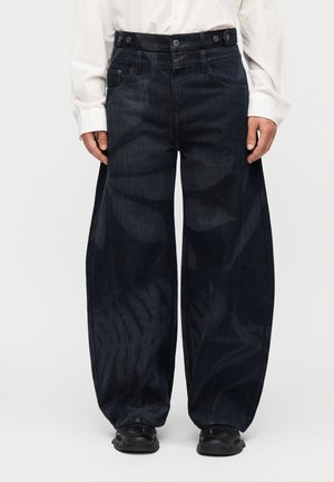 Άτομο που φοράει σκούρο ξεβαμμένο τζιν με φαρδιά μπατζάκια και διακριτικά αφηρημένα σχέδια, λευκό πουκάμισο και μαύρα αθλητικά παπούτσια σε απλό φόντο.