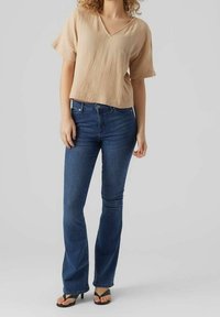 Haut beige texturé à col en V, associé à un jean évasé bleu. Le mannequin porte des sandales noires plates. Design simple avec une coupe décontractée et une taille mi-haute.