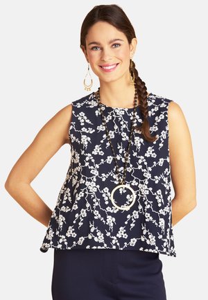 Lächelnde Frau mit ärmellosem marineblauem Oberteil mit Blumenmuster, langer Perlenkette mit rundem Anhänger, goldenen hängenden Ohrringen und geflochtenem Haar.