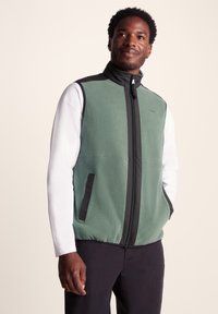 Fleece vest in mint green with black accents, full front zipper, two side pockets, and a high collar. Smooth texture and lightweight design.