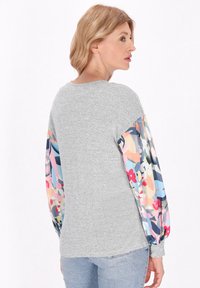 Szürke kapucnis pulóver színes virágmintás ujjakra, kerek nyakkivágással és lazább fazonnal. Az anyag puha és texturált.