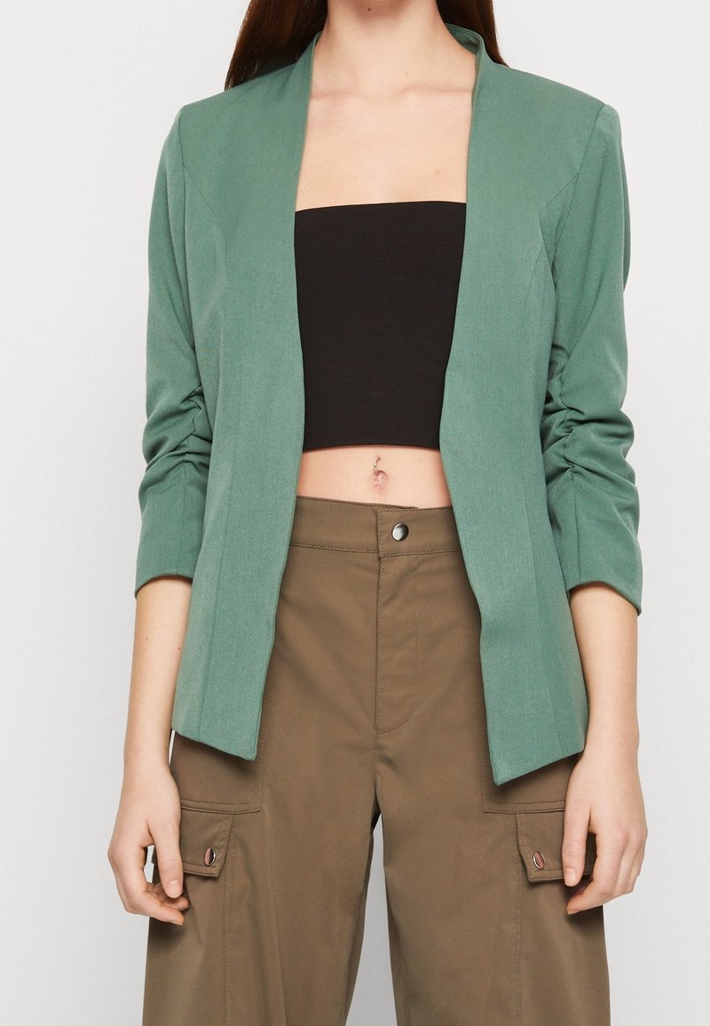 Femme portant un blazer vert par-dessus un crop top noir et un pantalon cargo marron, les mains détendues le long du corps, montrant le ventre et un anneau de nombril.
