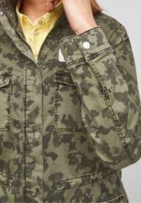 Persoon die de kraag van een groene en donkere camouflagejas met metalen knopen over een lichtgele shirt aanpast.