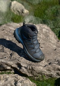 Tamsiai mėlyni žygio batai su juodais ir pilkais akcentais, ilsintis ant didelio akmens lauke su žalia veja fone.