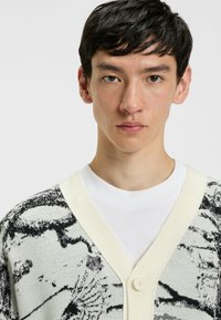 Strikket cardigan i sort og hvid med abstrakt mønster, der har en kridtfarvet V-udskæring og enkelt knaplukning.